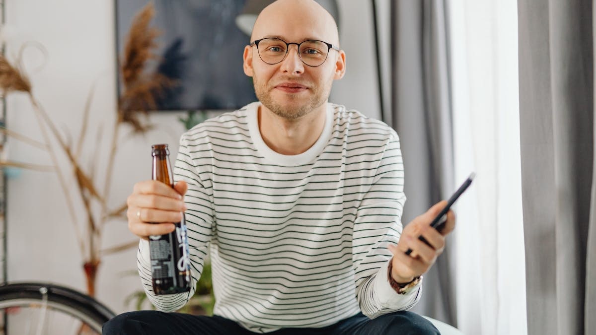 A cheerful man holding a beer bottle and smartphone, enjoying leisure time indoors.