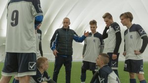 A coach passionately instructs his soccer team during a practice session inside a sports dome.